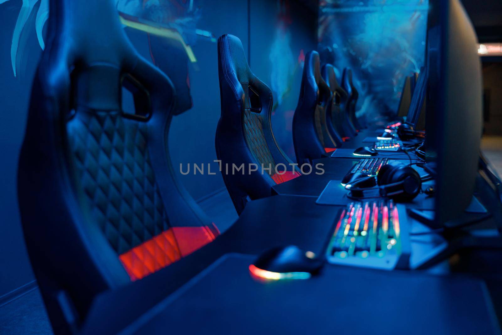 View on tables equipped with PC monitors, brightly lit keyboards and headsets with empty streaming chairs in dark gaming room