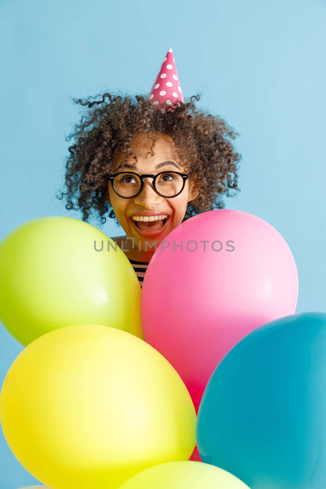 Cheerful multiethnic lady in birthday cone hat holding party balloons. Isolated on blue background