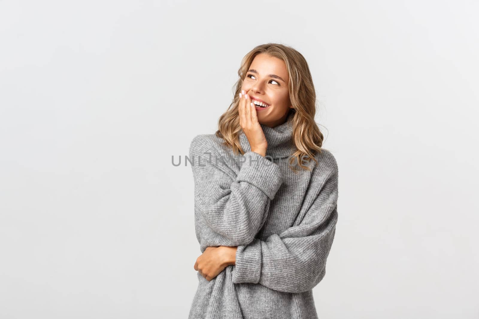 Portrait of attractive caucasian woman in grey sweater looking at logo at upper left corner, smiling and laughing, standing happy over white background.