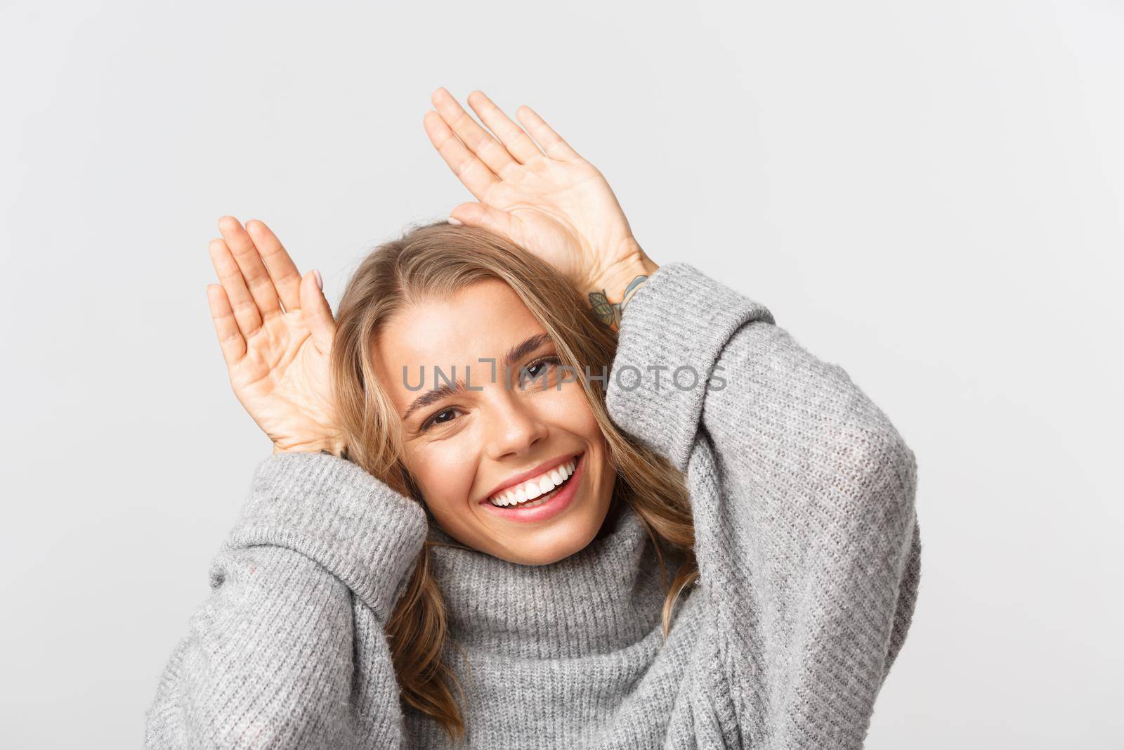 Close-up of lovely blond girl in grey sweater, smiling and showing puppy ears with hands raised over head, standing over white background.