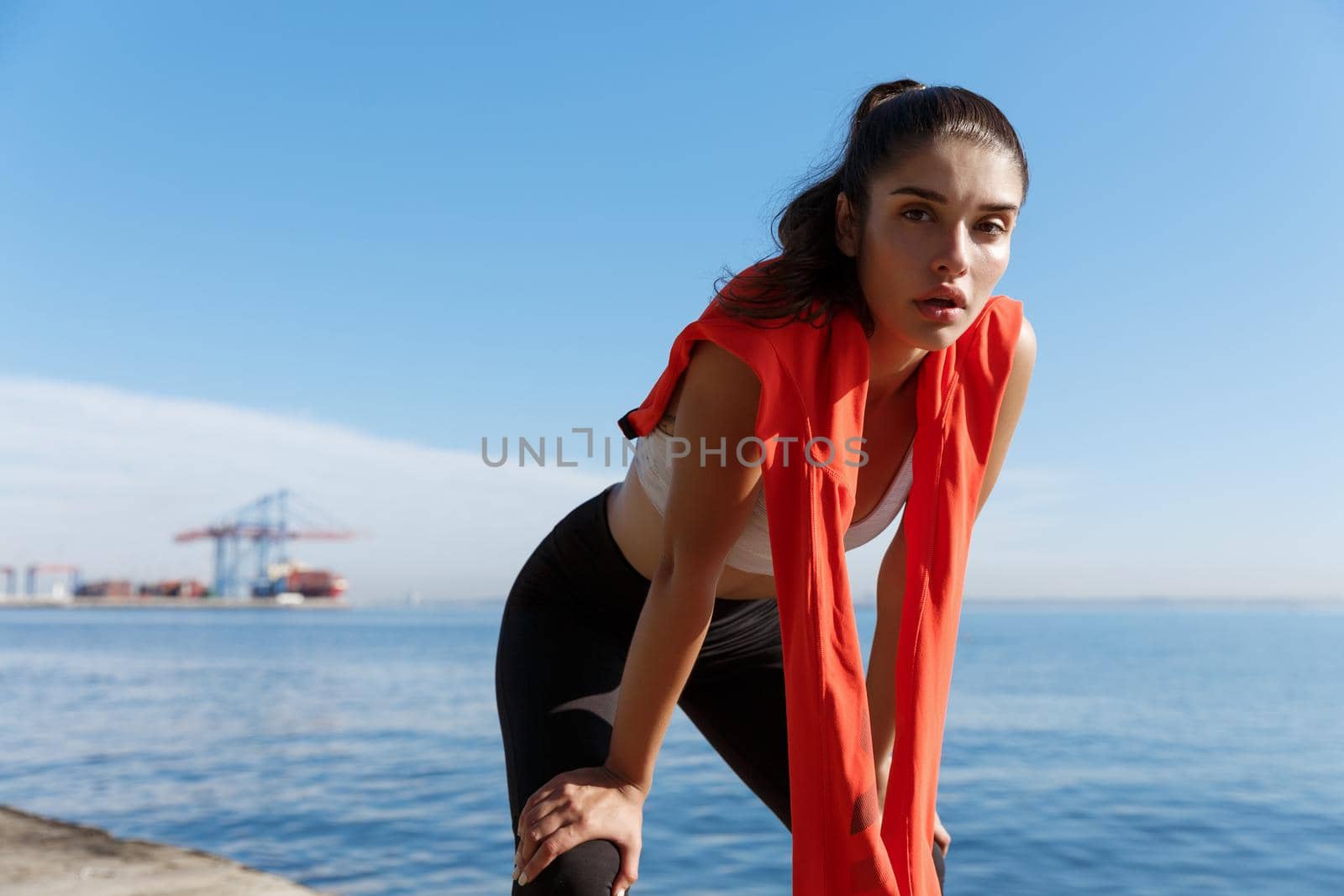 Outdoor shot of attractive sportswoman having a break after running on the seaside promenade. Woman taking a breath and panting during jogging.