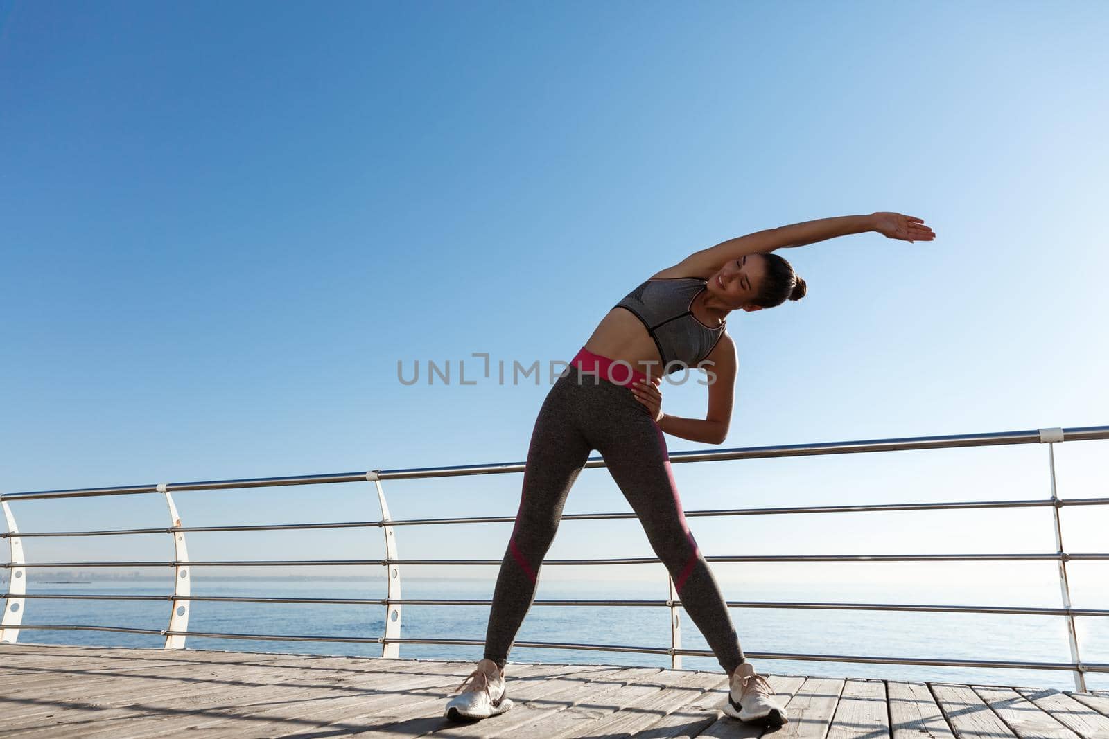 Outdoor shot of attractive fitness woman training on a pier. Girl stretching before workout, standing on the seaside promenade by Benzoix