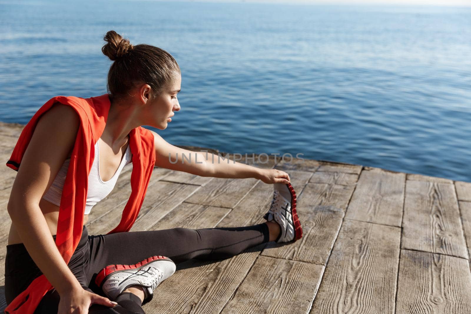 Outdoor shot of attractive fitness woman warming-up before jogging, sitting on pier and stretching legs.
