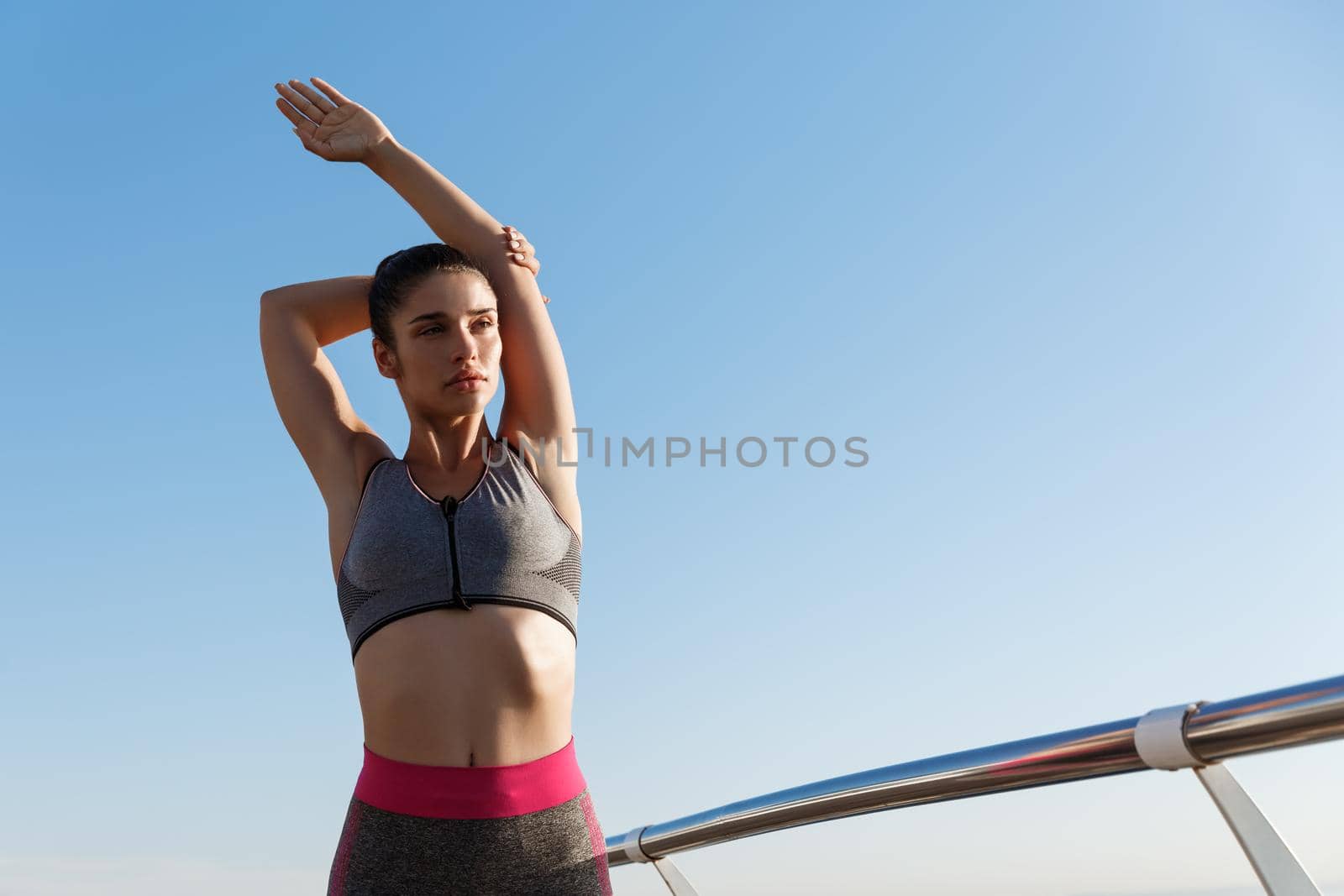 Young attractive sportswoman stretching hands and warming-up before jogging on the seaside promenade. Female athlete training outdoors on a pier.