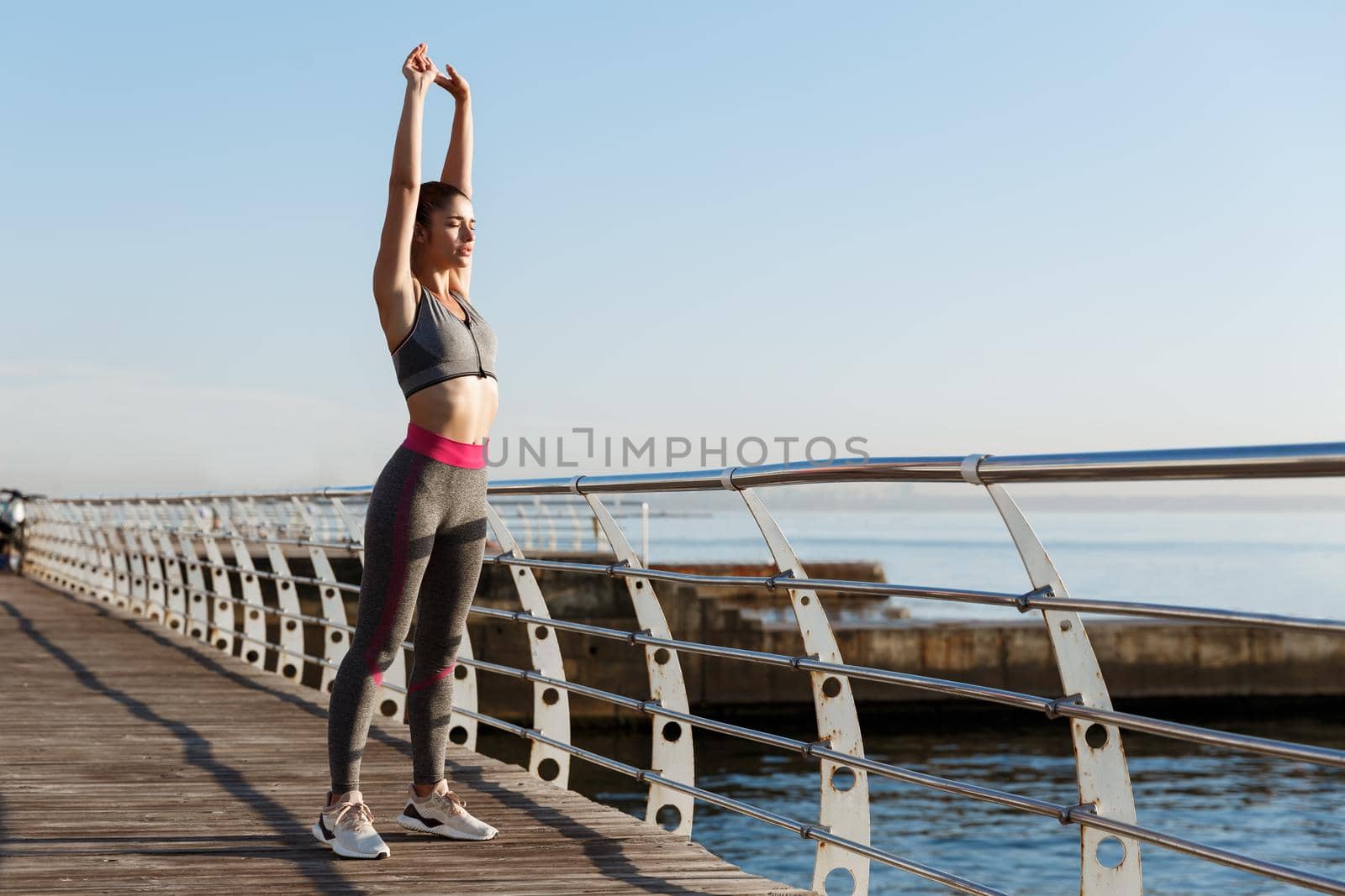 Young fit sportswoman stretching before morning exercises. Female runner workout on seaside promenade.