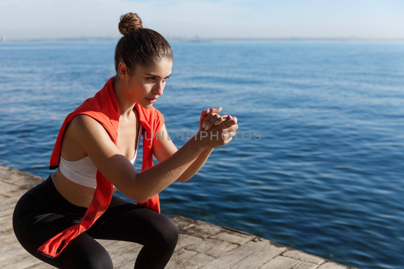 Side view of focused young sportswoman doing squats near the sea, having training on seaside by Benzoix