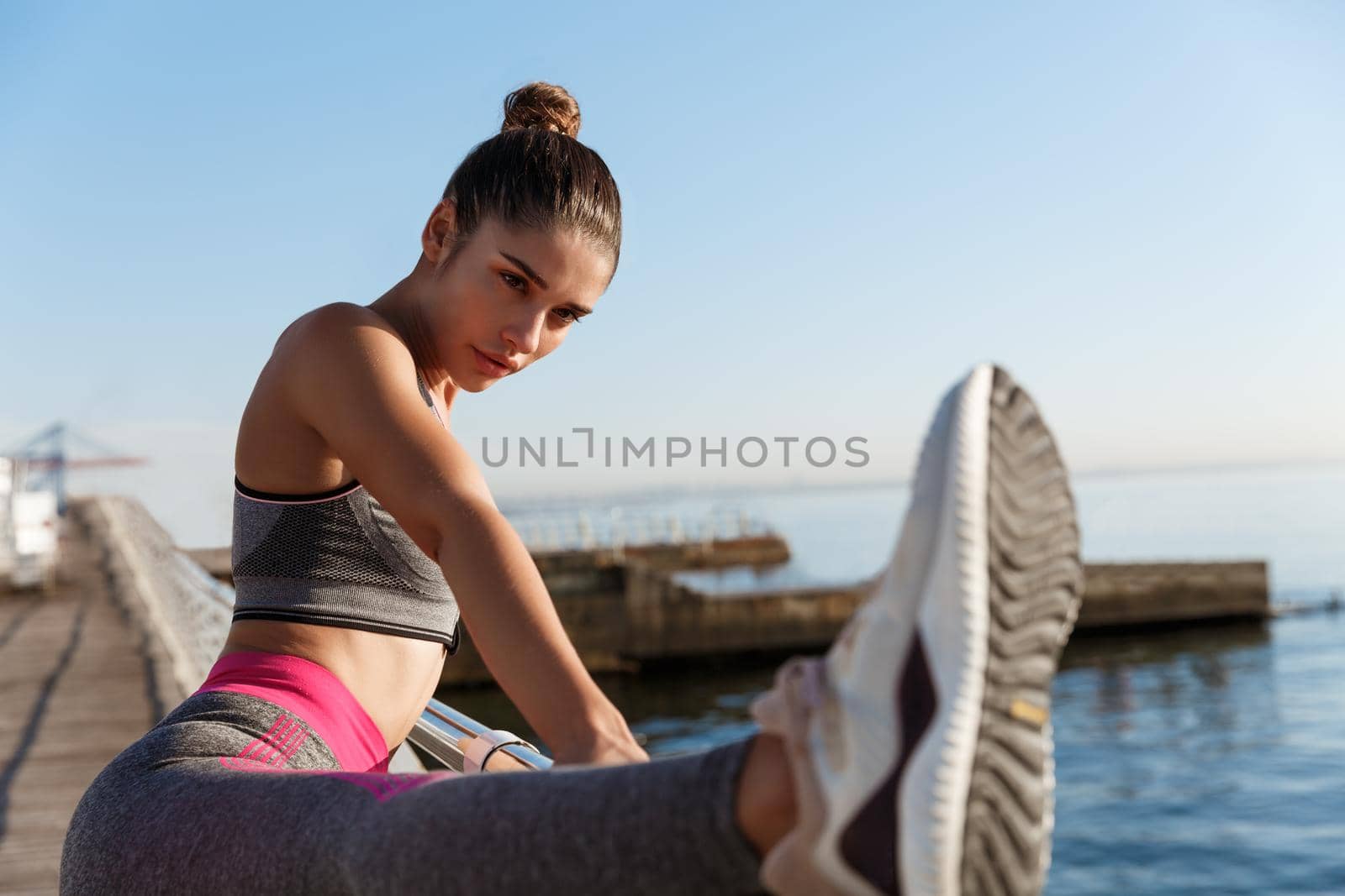 Close-up of attractive sportswoman holding leg on handrope, stretching on the seaside promenade. Female runner warming-up for morning jogging training by Benzoix