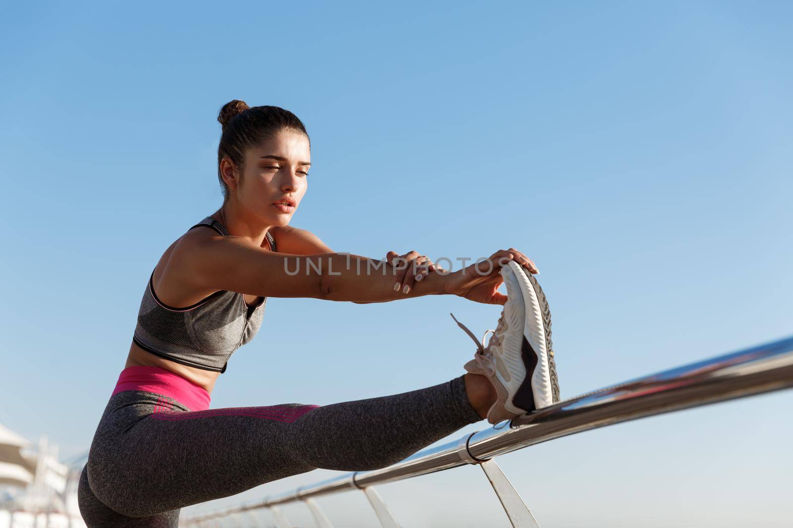 Outdoor shot of attractive healthy woman warming-up before training. Girl stretching legs on a pier, prepare for run along seaside.