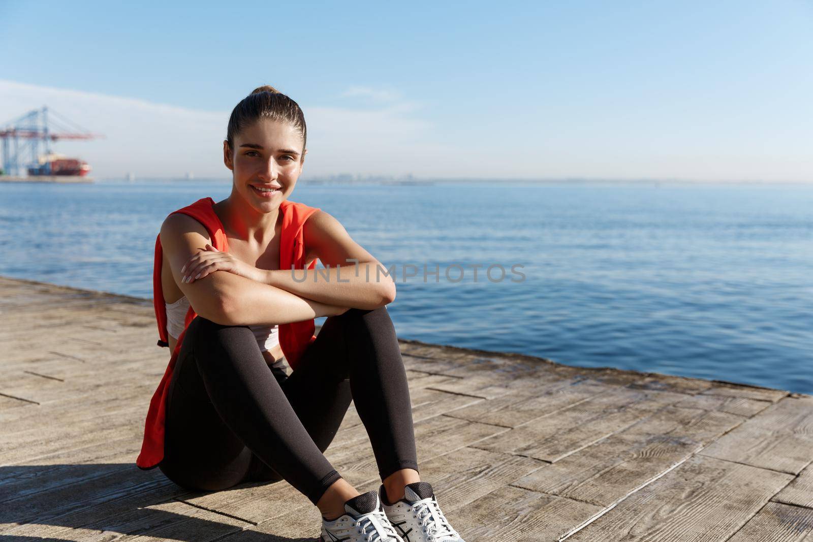 Outdoor shot of beautiful young sportswoman sitting on wooden pier and smiling. Fitness woman resting after workout near the sea, enjoying view.