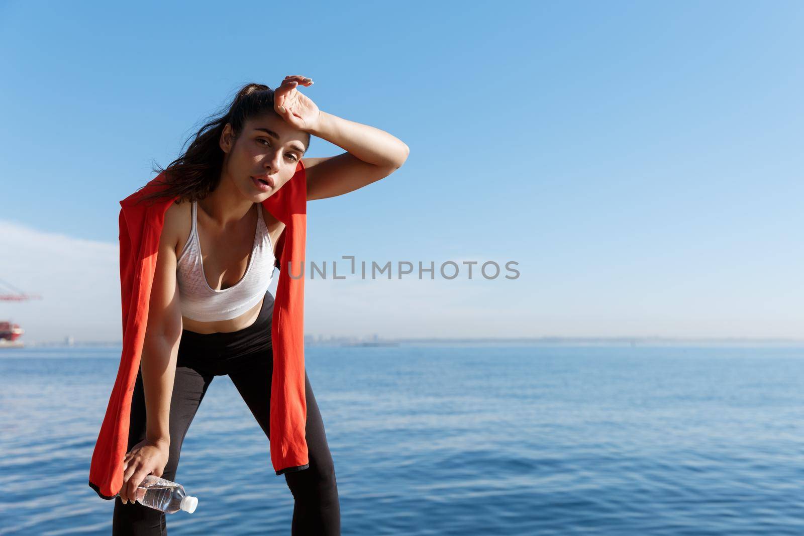 Outdoor shot of tired young sportswoman having a break near the sea, wiping sweat and panting after running.