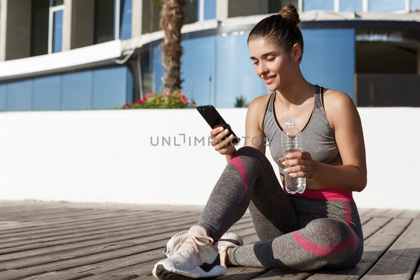 Outdoor shot of attractive sportswoman sitting on wooden pier with mobile phone, having break after workout, drinking water.