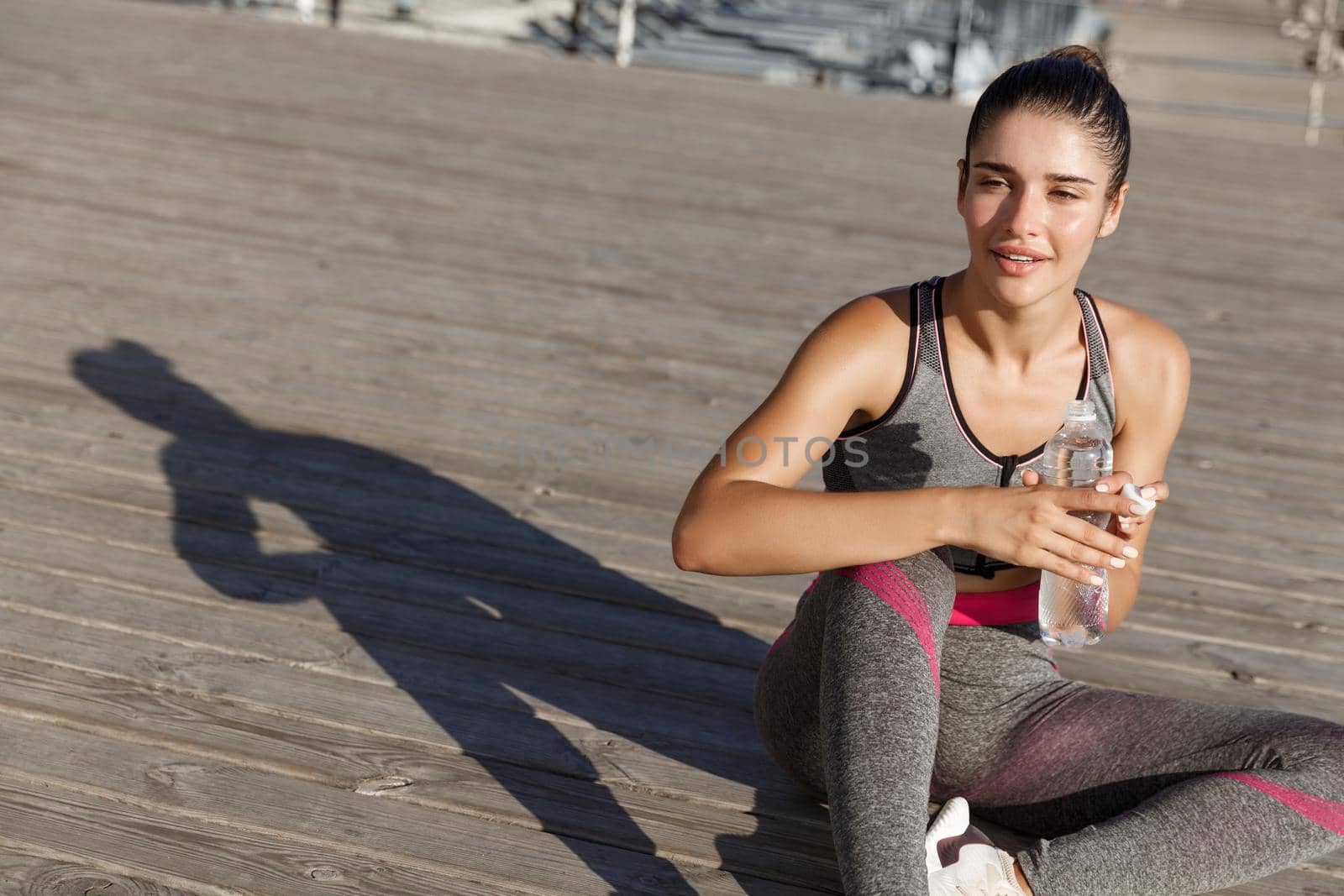 Close-up of attractive sportswoman taking a break after workout, sitting at the seaside and drinking water, looking pleased by Benzoix