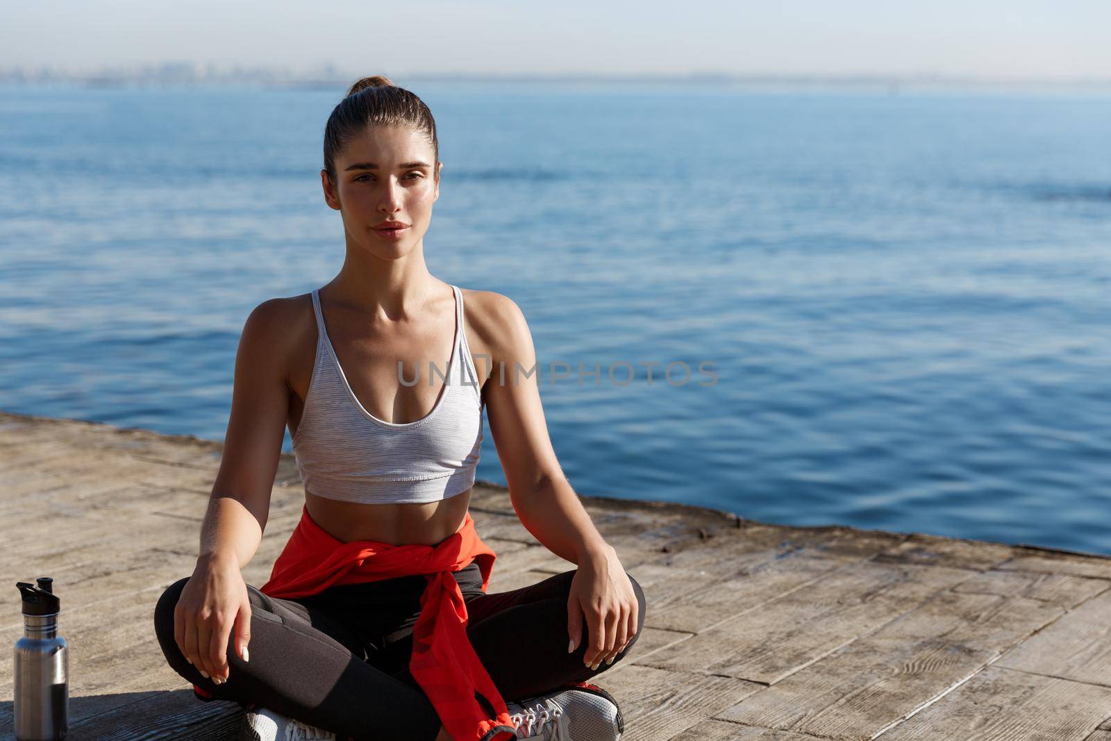 Outdoor shot of relaxed young woman meditating near the sea, practice yoga, sitting in asana on a wooden pier floor by Benzoix