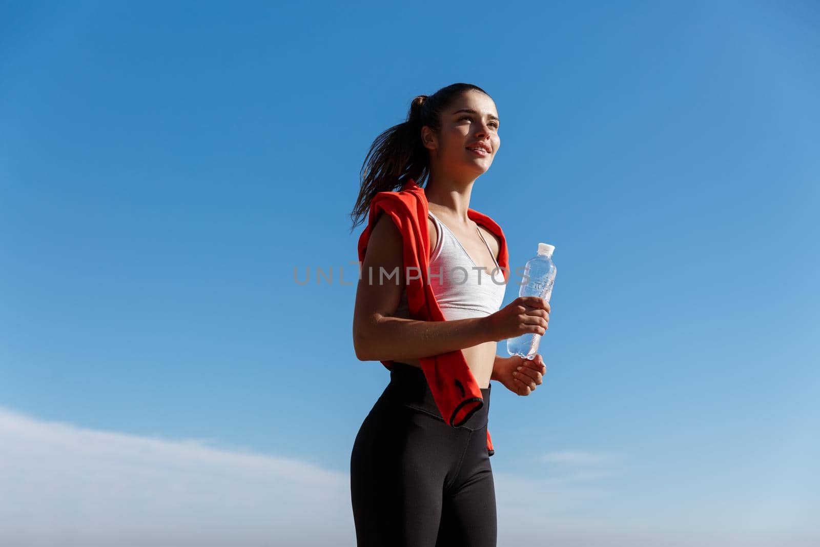 Low angle shot of happy and confident fitness woman jogging in the morning, running and smiling, holding bottle of water by Benzoix