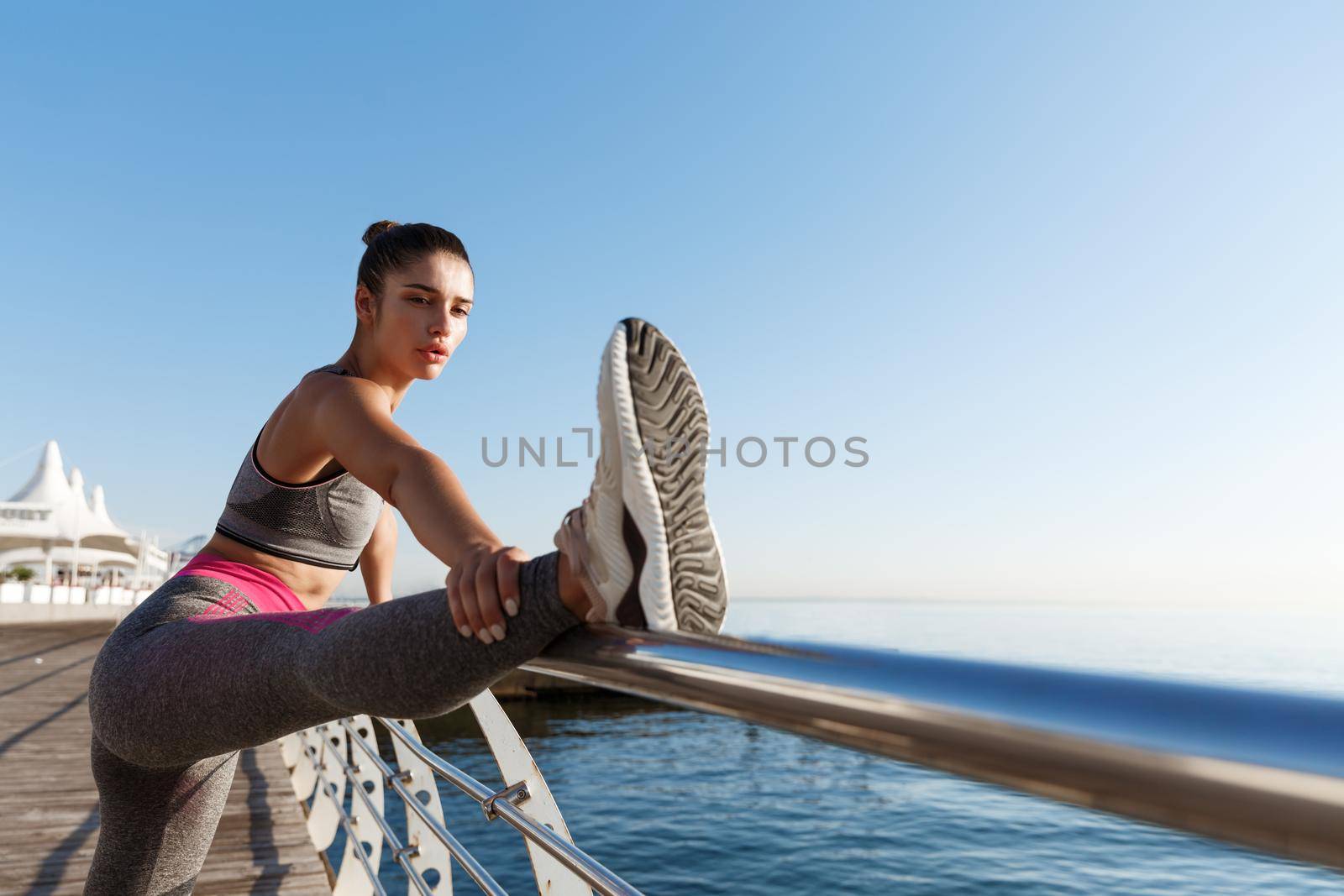 Outdoor shot of attractive healthy woman in sports clothing, stretching her legs and warming-up before jogging workout near sea by Benzoix