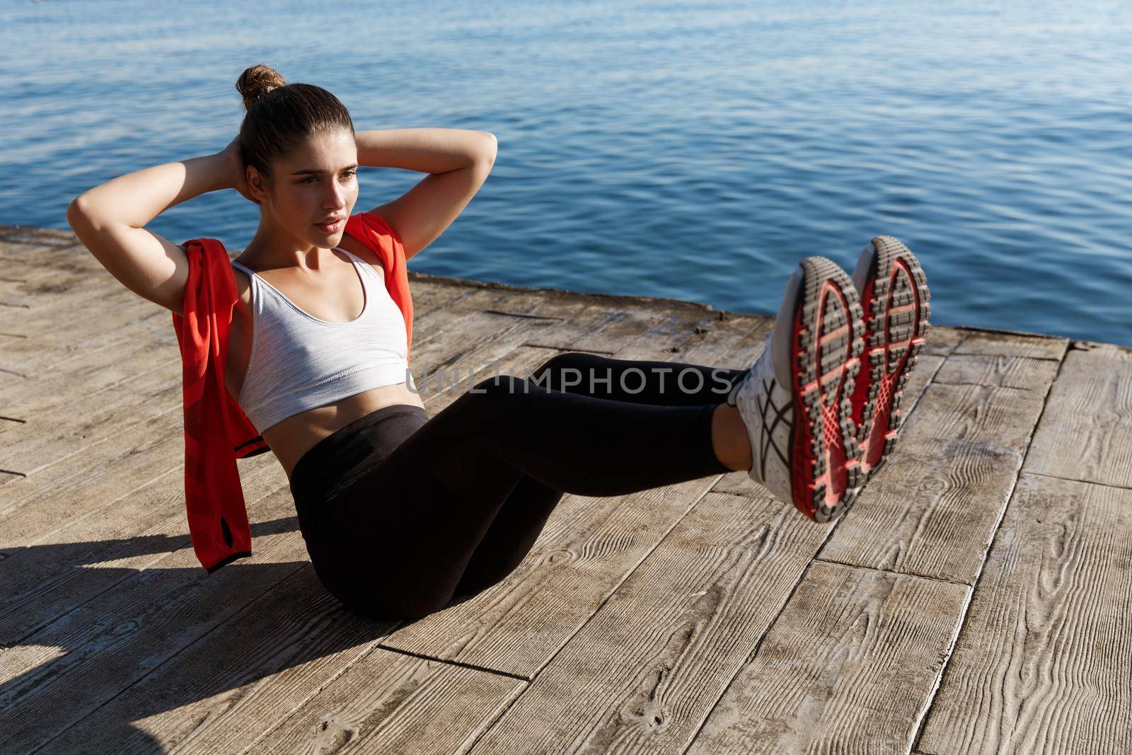 Outdoor shot of confident fitness woman working on her abs. Sportswoman doing crunches with raised legs, training near sea on a pier by Benzoix