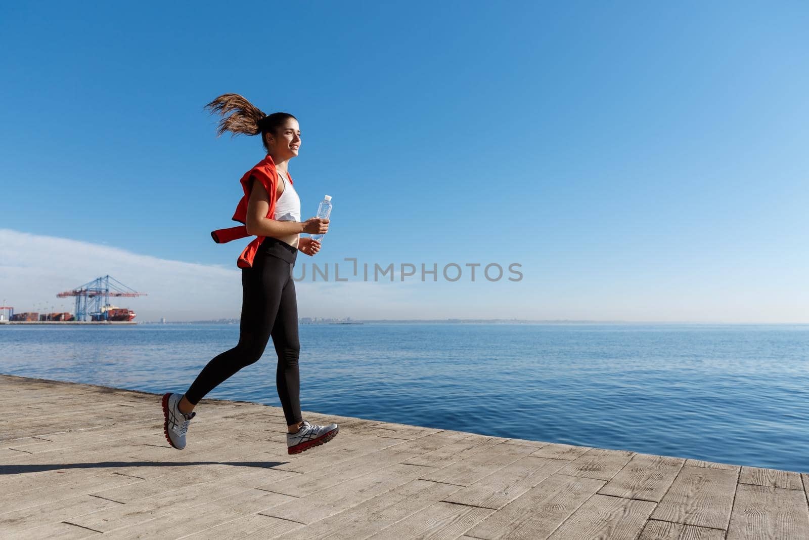 Side view of athletic young woman running along the seaside promenade. Sportswoman jogging near the sea with bottle of water by Benzoix