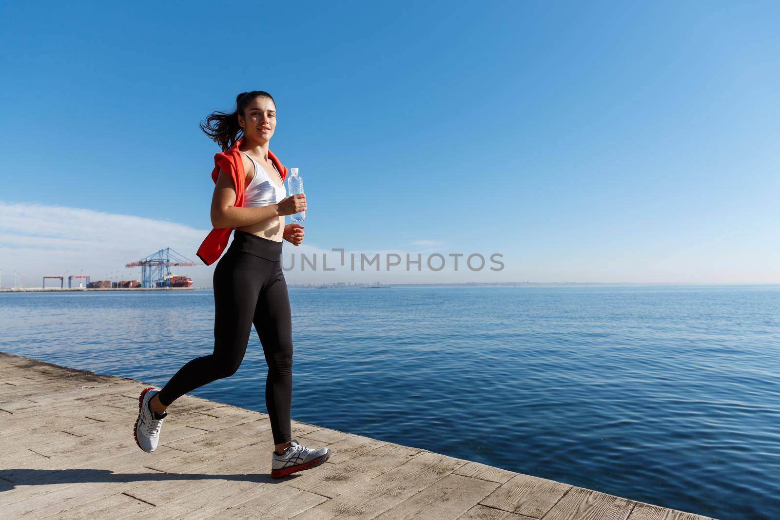 Outdoor shot of smiling sportswoman jogging and working out on seaside promenade. Fitness woman running and looking at camera by Benzoix