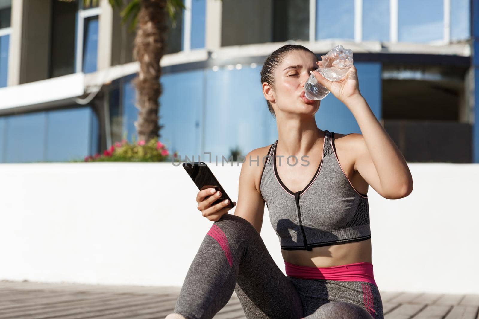 Outdoor shot of attractive fitness woman sitting on wooden pier with mobile phone, having break after workout, drinking water.
