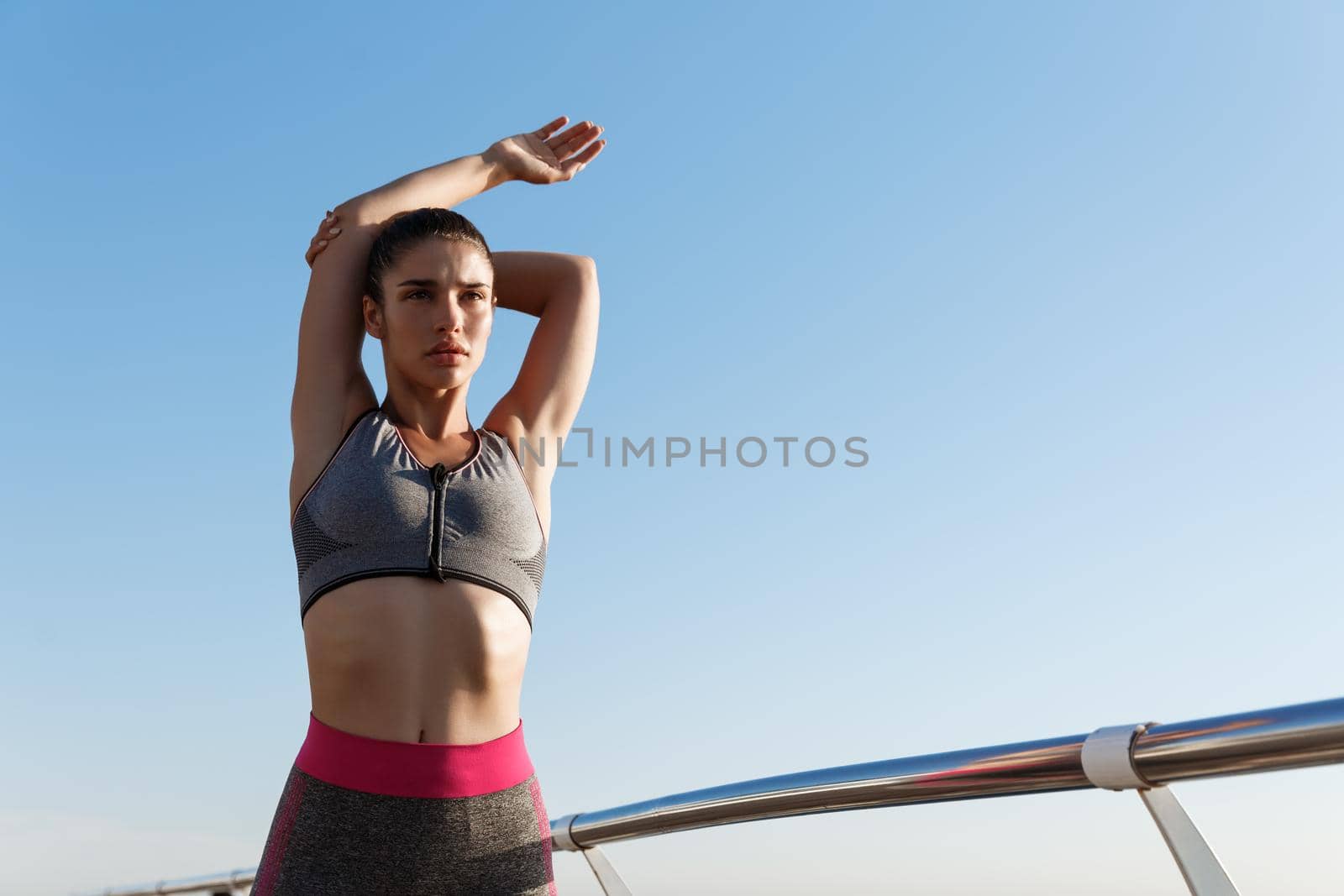 Low angle of attractive woman stretching and warming-up before jogging workout, looking determined forward, standing on a pier with sky behind her.
