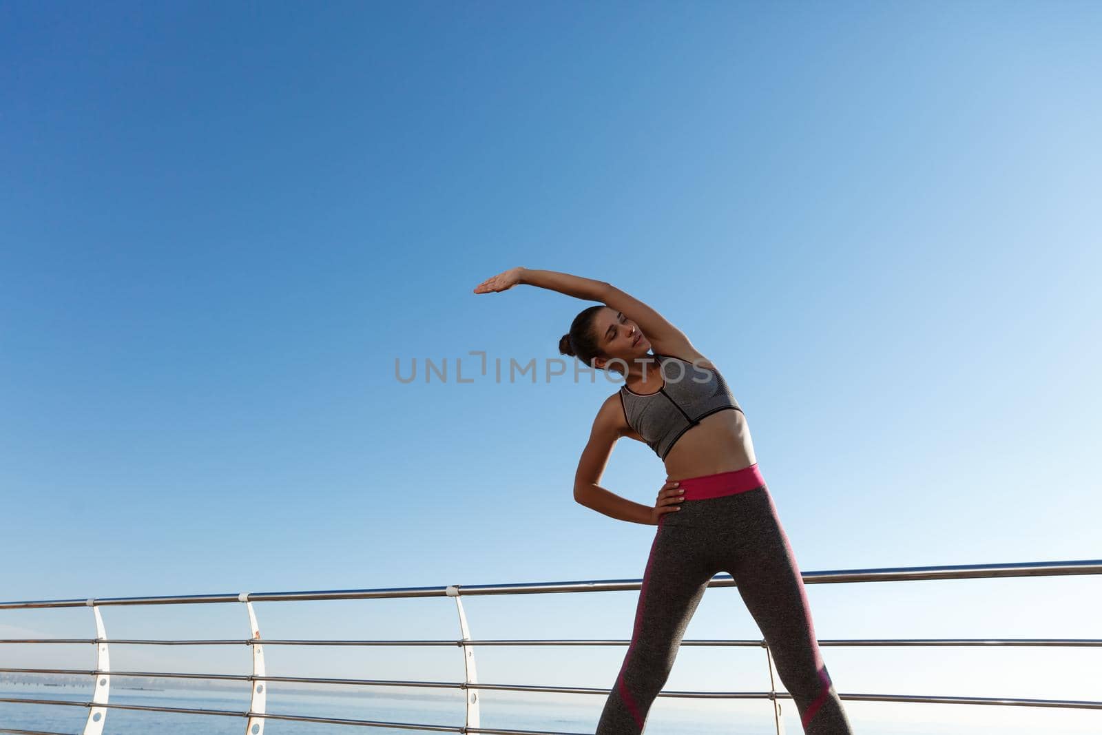 Image of attractive healthy woman stretching and training on a pier. Girl having workout on fresh air.