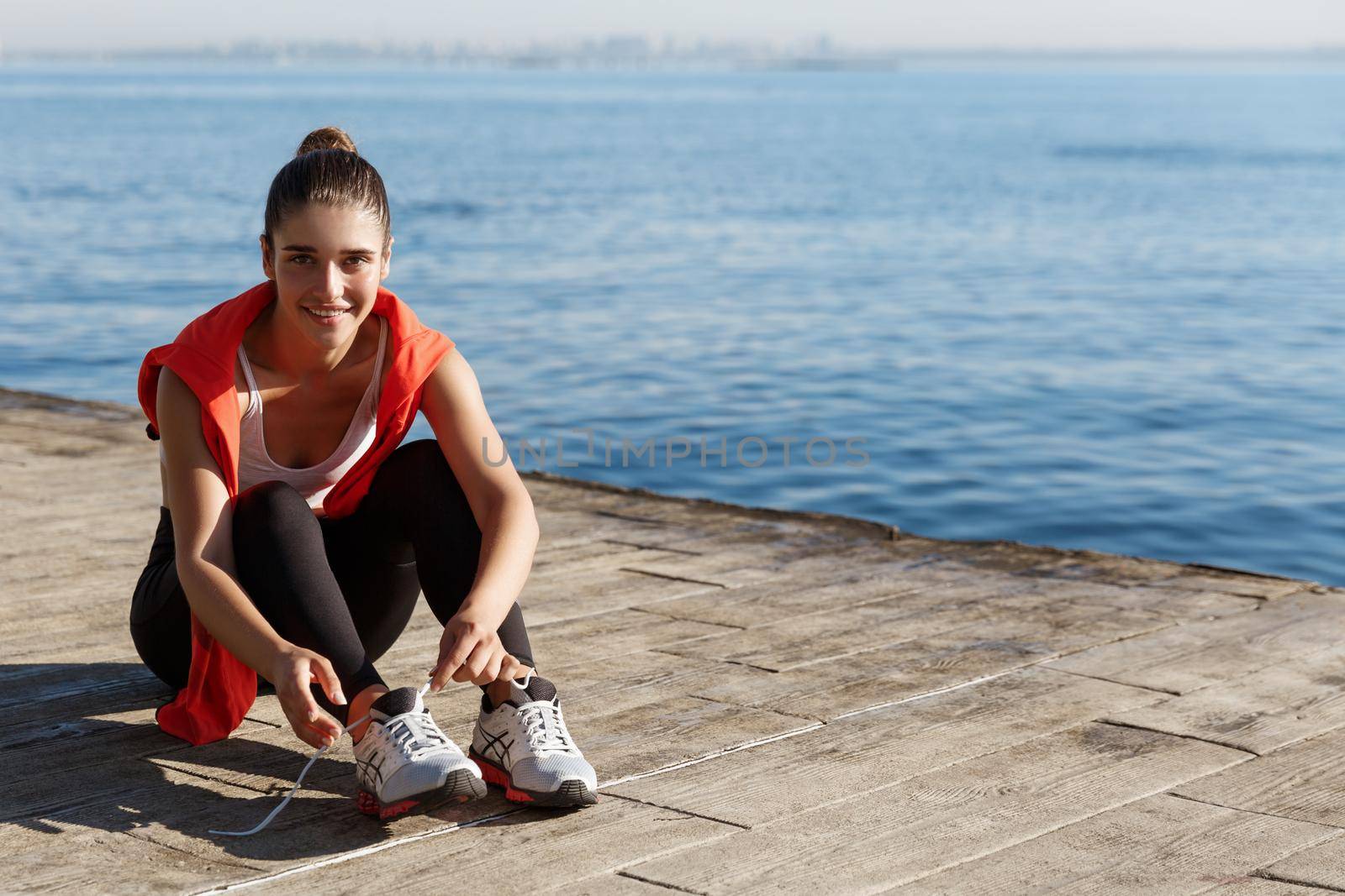 Outdoor shot of attractive smiling sportswoman sitting on pier and tying shoelaces during workout, training near the sea.