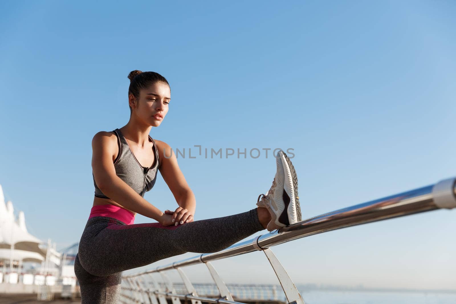 Image of attractive fit fitness girl workout on a pier. Sportswoman stretching legs and warming-up before jogging training on the seaside promenade.