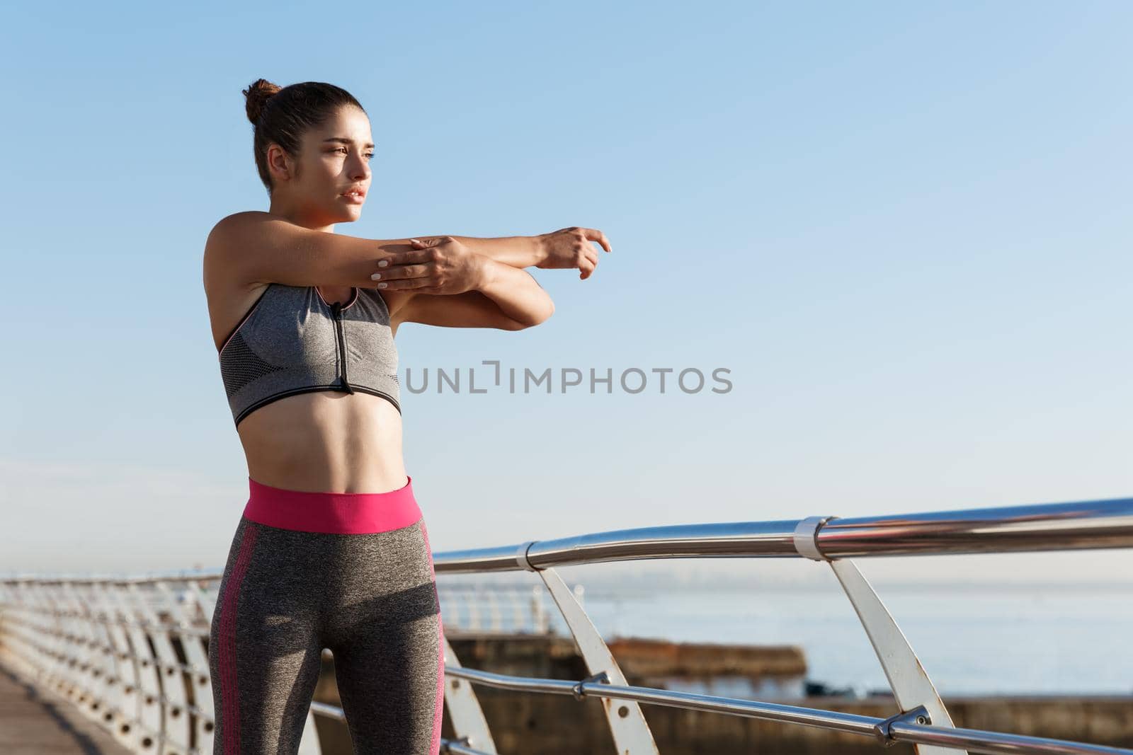 Image of determined female runner stretching on the seaside promenade before jogging. Girl warming-up on pier to start workout, looking at the sea by Benzoix