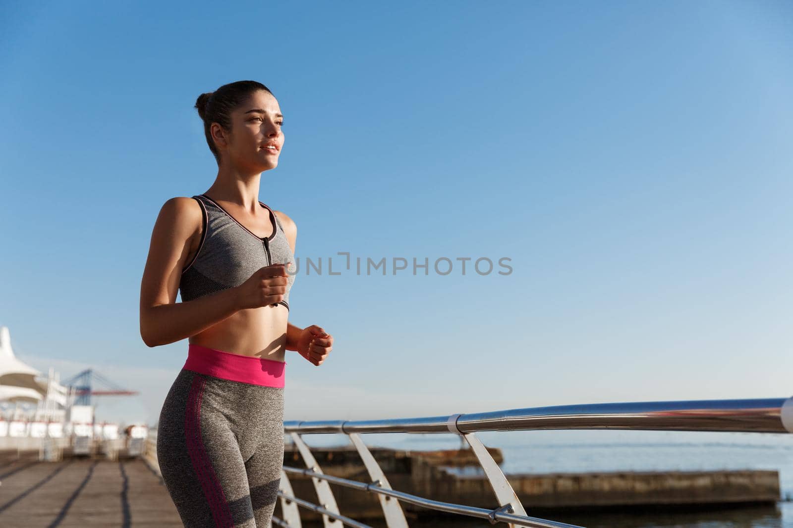Image of young attractive sportswoman workout alone near the sea. Female athlete jogging on a pier.