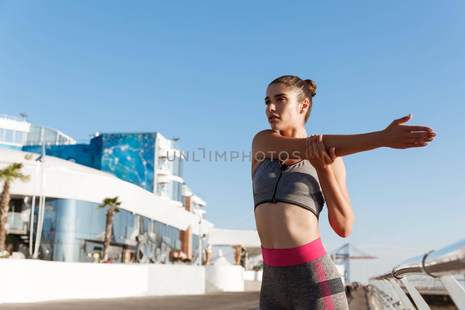 Young attractive woman stretching hands and warming-up before workout. Runner doing exercises on the seaside pier, preparing for jogging.