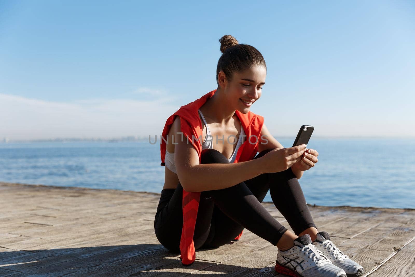 Outdoor shot of happy fitness woman sitting on wooden pier and smiling, using smartphone, having break after workout near the sea.