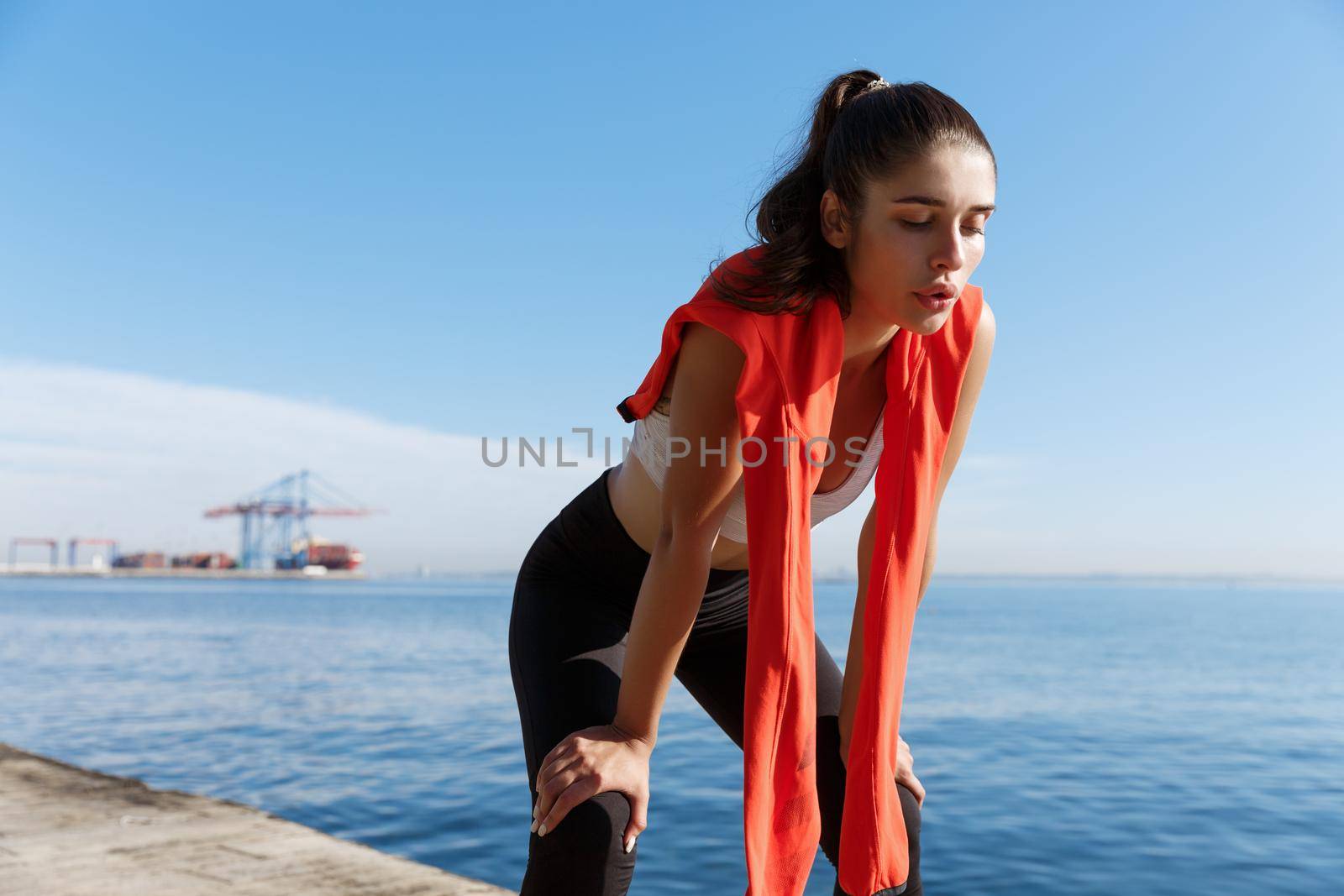 Outdoor shot of tired fitness woman panting and taking a breath after jogging, standing on a pier with sea behind her.