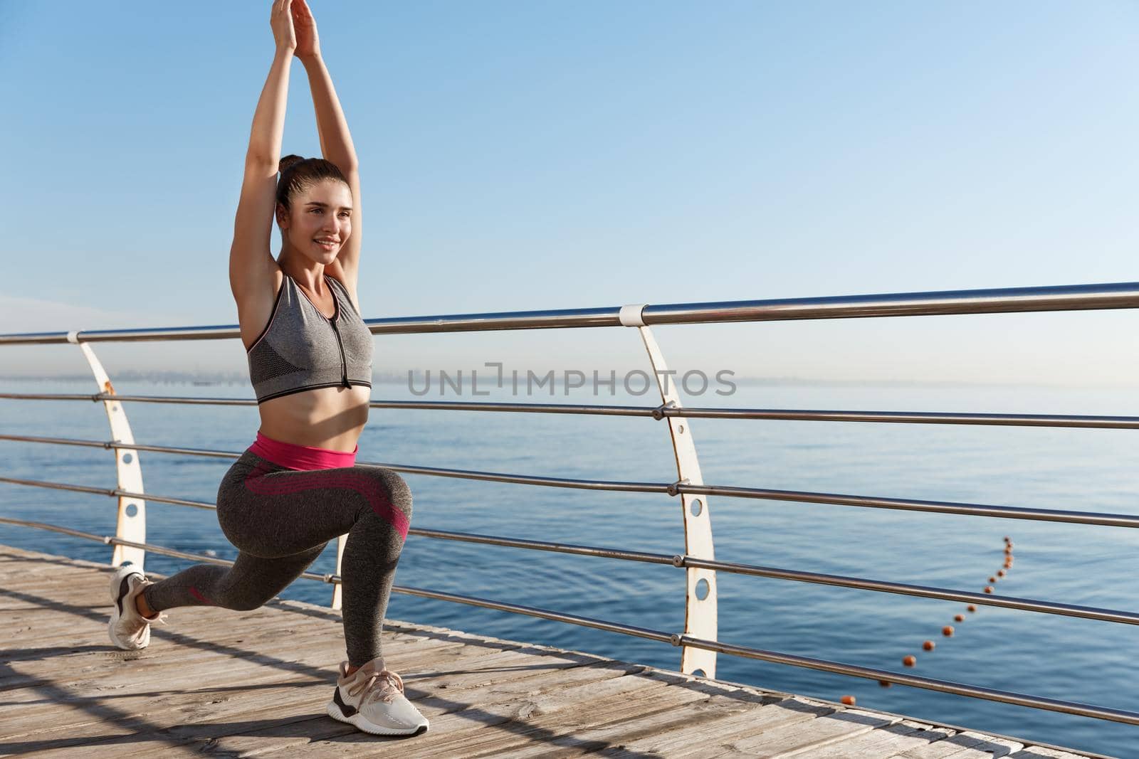 Outdoor shot of attractive young fitness woman stretching, warming-up before workout, doing exercises at the seaside.