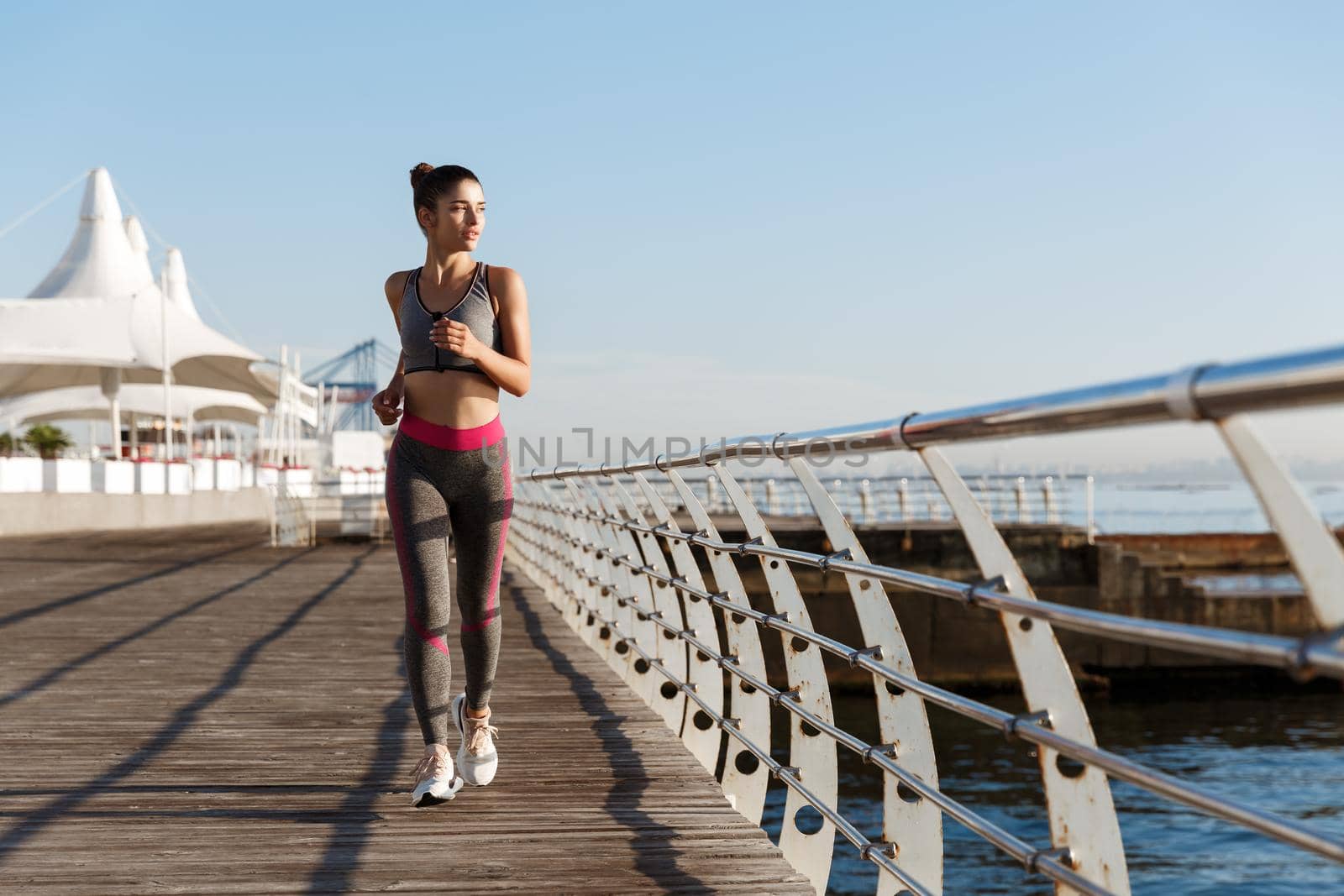 Full length shot of attractive young woman running along seaside promenade towards camera, looking at the sea. Female runner training in morning on a pier.