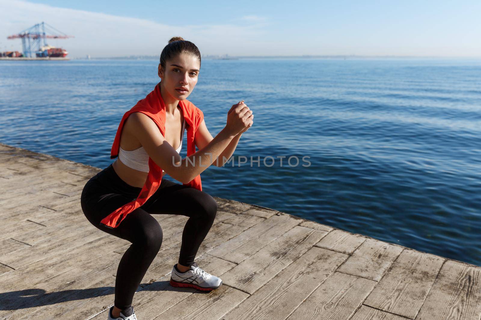 Outdoor shot of attractive fitness woman workout near the sea, doing squats exercises and looking at camera.