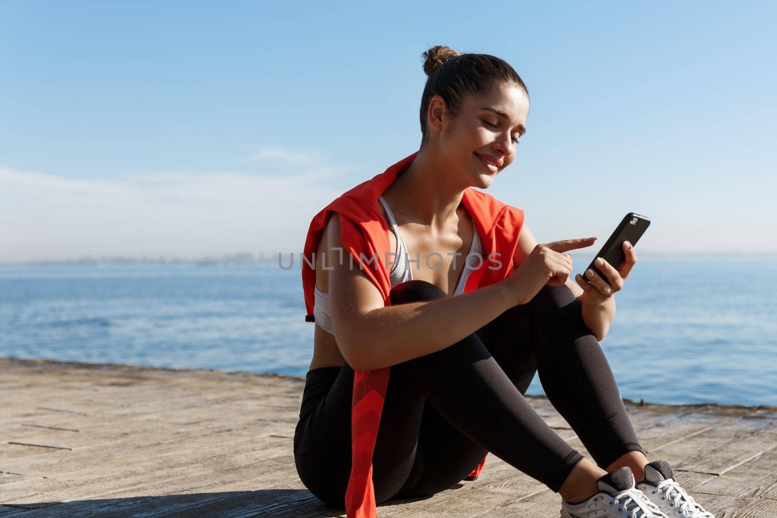 Outdoor shot of attractive sportswoman having a break near sea, sitting on wooden pier and using mobile phone.