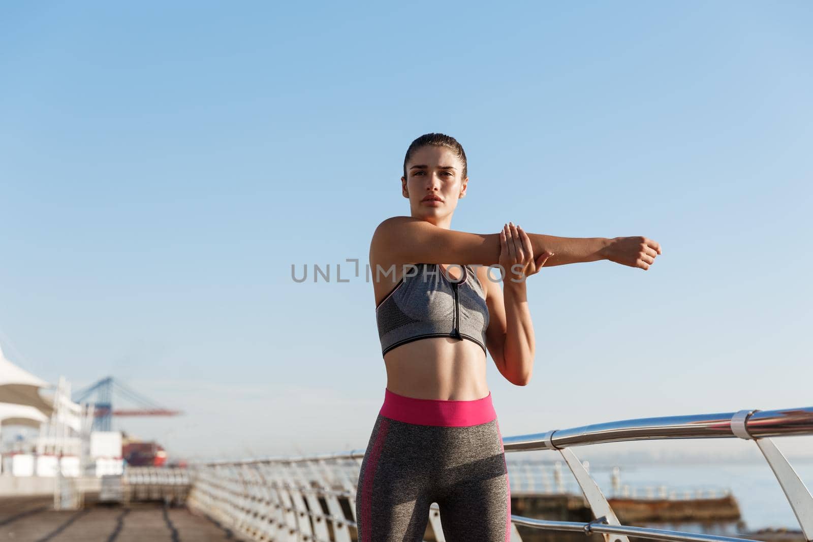 Image of beautiful healthy woman in fitness clothing stretching her body before jogging training. Female runner standing on the seaside promenade, training near the sea.