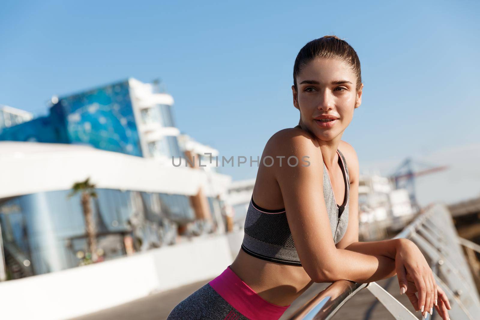 Close-up of good-looking healthy woman standing on a pier, leaning on handbar and looking aside, taking a breath during workout or jogging at seaside by Benzoix