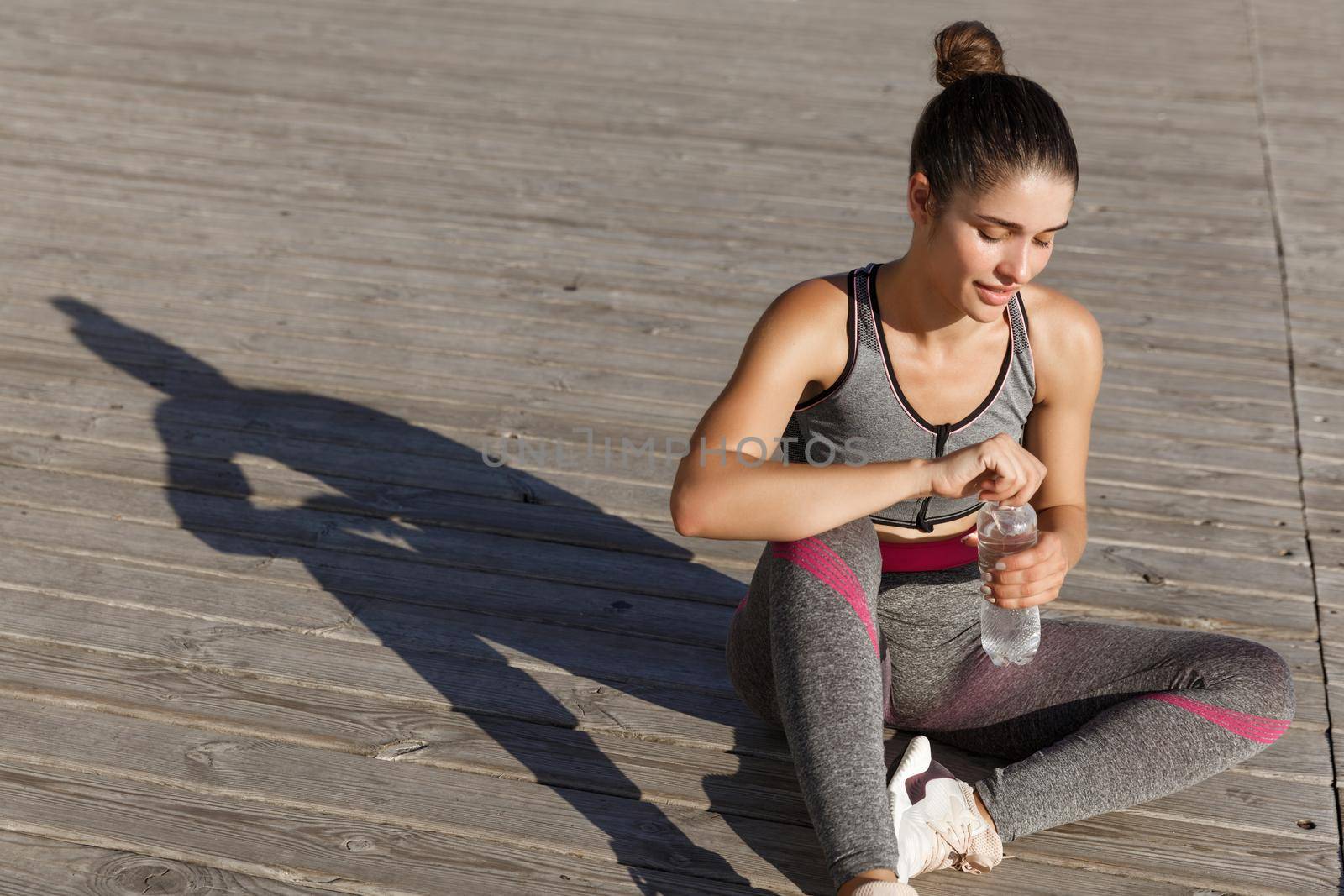 Outdoor shot of attractive fitness woman having a break after jogging, sitting on floor and drinking water, working out near the sea.