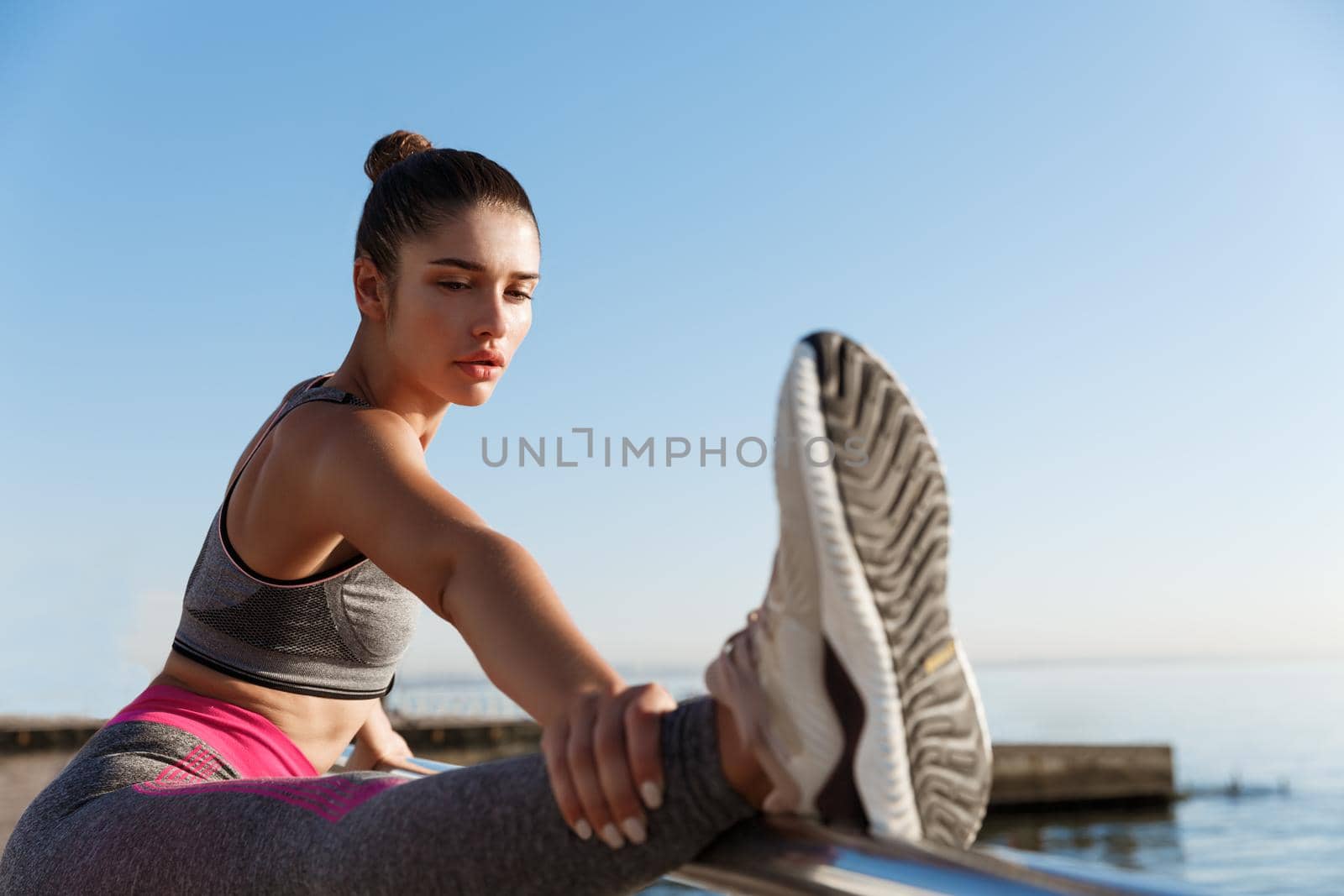Close-up of attractive sportswoman holding leg on handrope, stretching on the seaside promenade. Female runner warming-up for morning run and workout by Benzoix