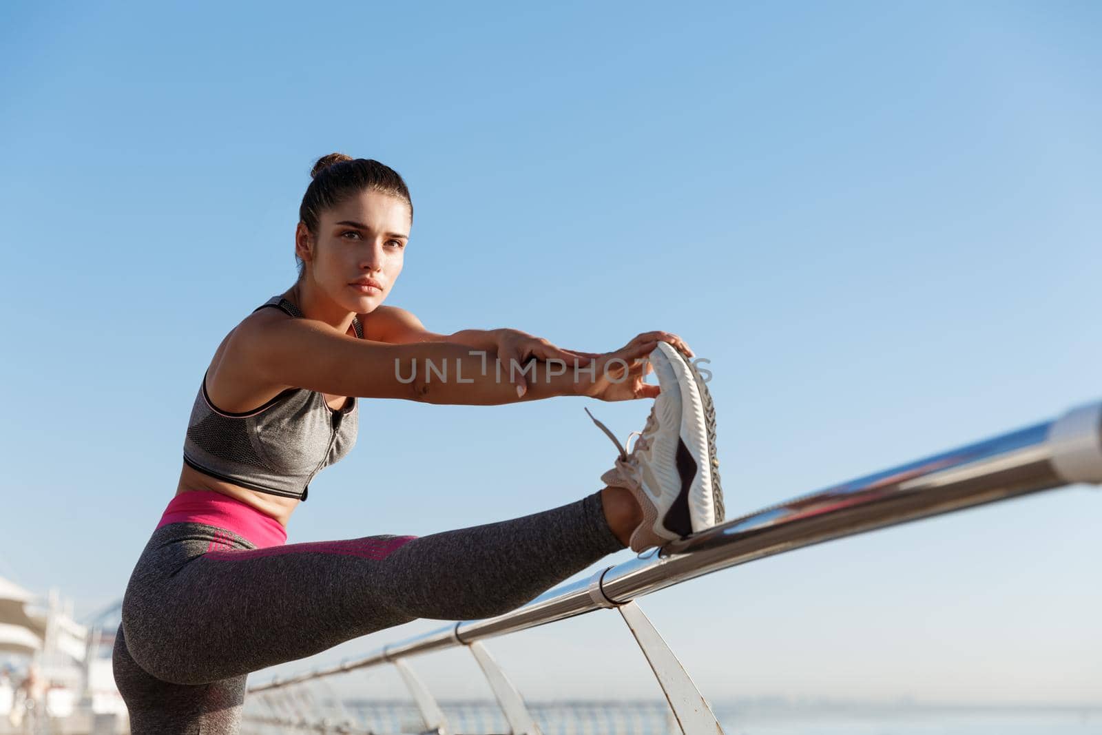 Determined sportswoman stretching leg on a pier and looking along the seaside promenade, warming-up before jogging workout by Benzoix