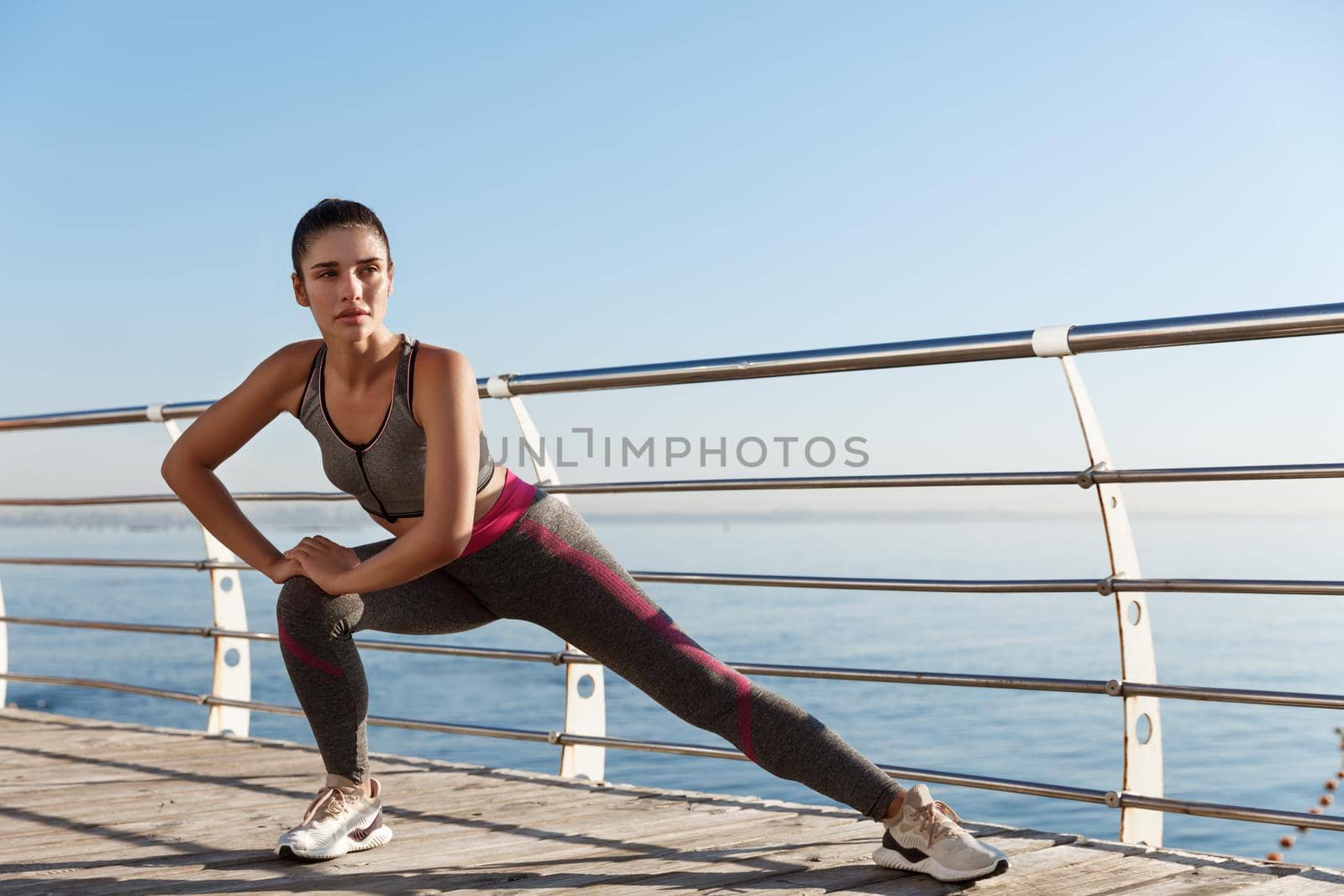 Outdoor shot of attractive and healthy fitness woman training near the sea, stretching legs before jogging.