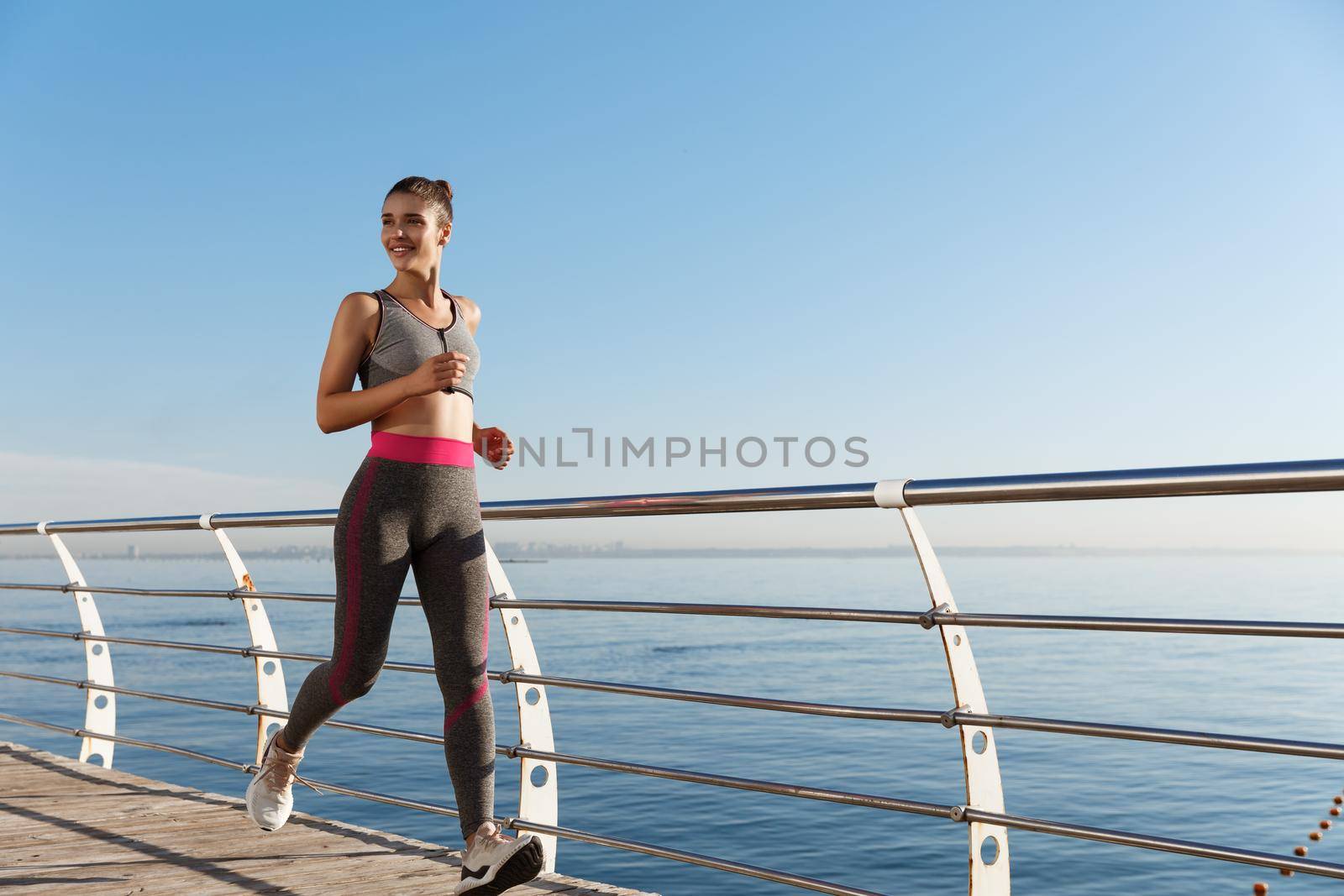 Outdoor shot of happy fitness woman running along seaside and smiling by Benzoix