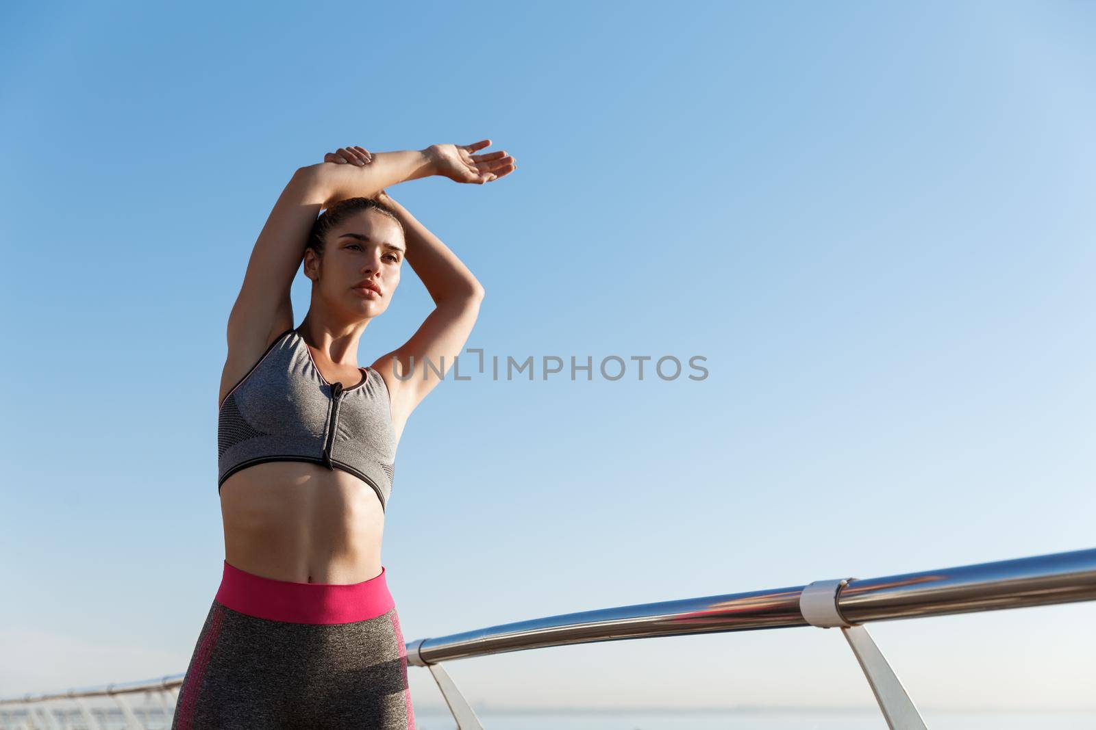 Low angle shot of attractive fit woman in sports bra and leggings, doing stretch exercises before jogging. Girl workout on the seaside promenade, prepare to run marathon by Benzoix