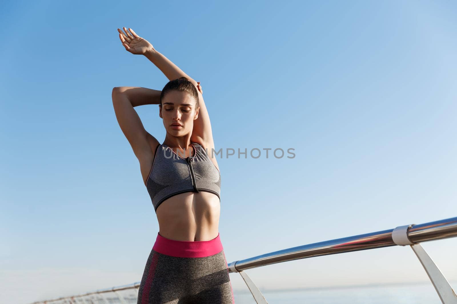 Image of young attractive woman stretching body before running workout at the seaside promenade. Sportswoman warm-up for jogging and training on pier.
