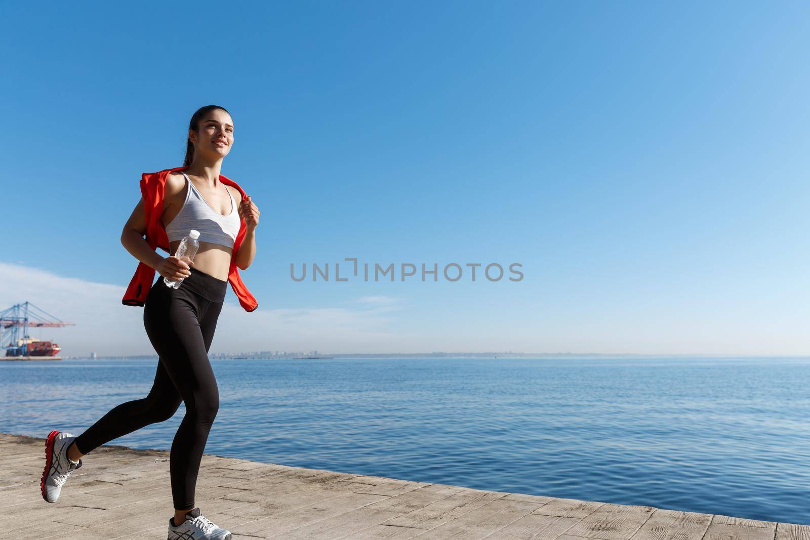 Outdoor shot of smiling sportswoman running along seaside and enjoying the view. Female athlete jogging near the sea.