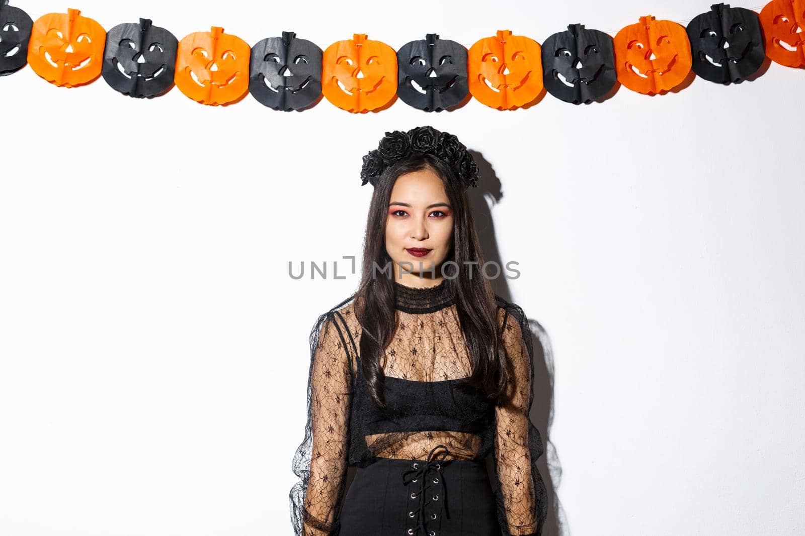 Portrait of elegant asian woman in witch costume, looking confident and standing against pumpkin streamers, decorations for halloween over white background by Benzoix