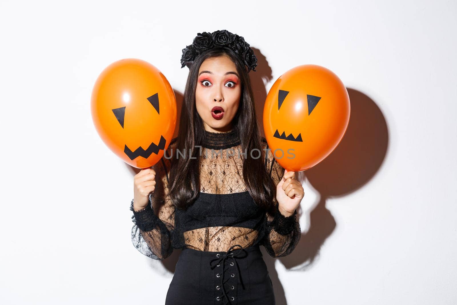 Image of surprised asian woman in witch costume celebrating halloween, holding balloons with scary faces, standing over white background by Benzoix