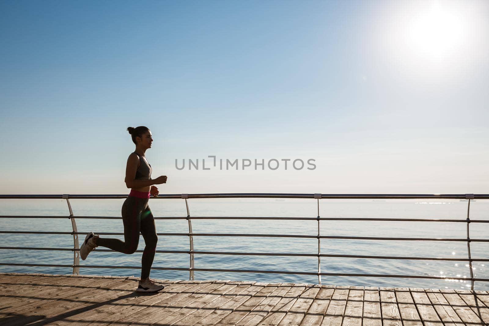 Side view of young fitness woman running along pier. Female athlete workout and jogging on the seaside promenade.