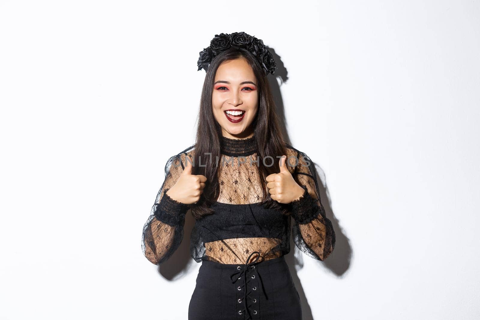 Image of happy satisfied asian woman dressed-up for halloween party, showing thumbs-up in approval, smiling pleased over white background. Girl in witch costume looking delighted.